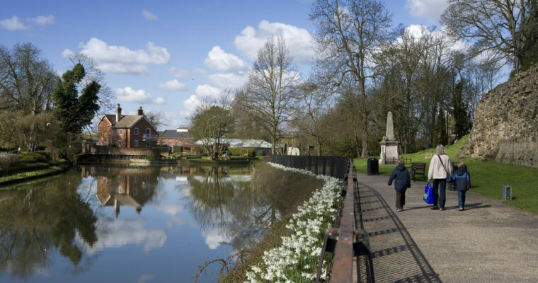 A woman and two children walking along a canal path. There are clouds in a blue sky and you can see the reflections of the clouds and trees in the canal