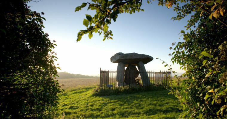 An ancient stone monument known as Kit’s Coty House, featuring large, upright sarsen stones arranged in a rectangular formation, set against a backdrop of lush green fields and a clear blue sky.