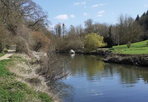 A calm river with a small boat in the distance. Grass and trees are on either side and a blue cloudy sky.