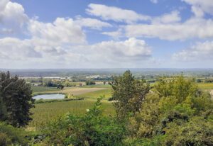 A scenic countryside landscape with rolling green fields, clusters of trees, and a small pond in the distance beneath a bright sky filled with white clouds