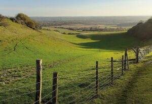 A bright green valley surrounded by gentle hills, with a wire fence running along the right side and sweeping views of farmland in the distance.
