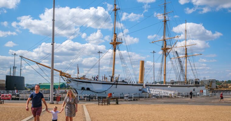 A family walking on a pathway in front of a historic ship named HMS Gannet, docked under a blue sky with scattered clouds