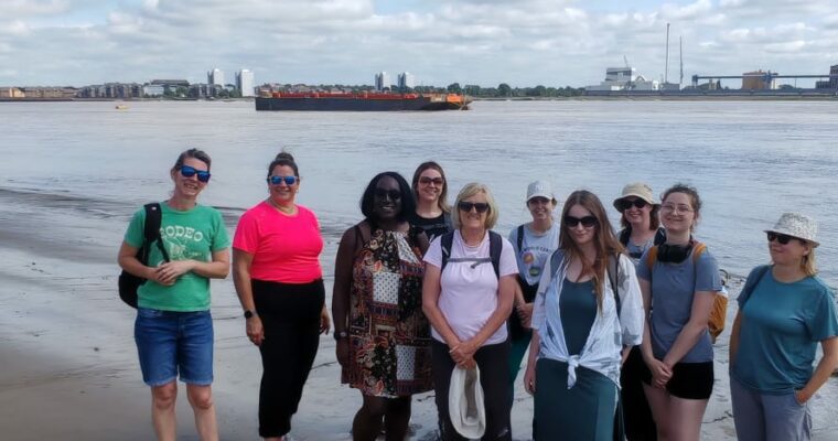 Group of women smiling at the camera standing on Northfleet beach. The River Thames is in the background with a boat in the background.