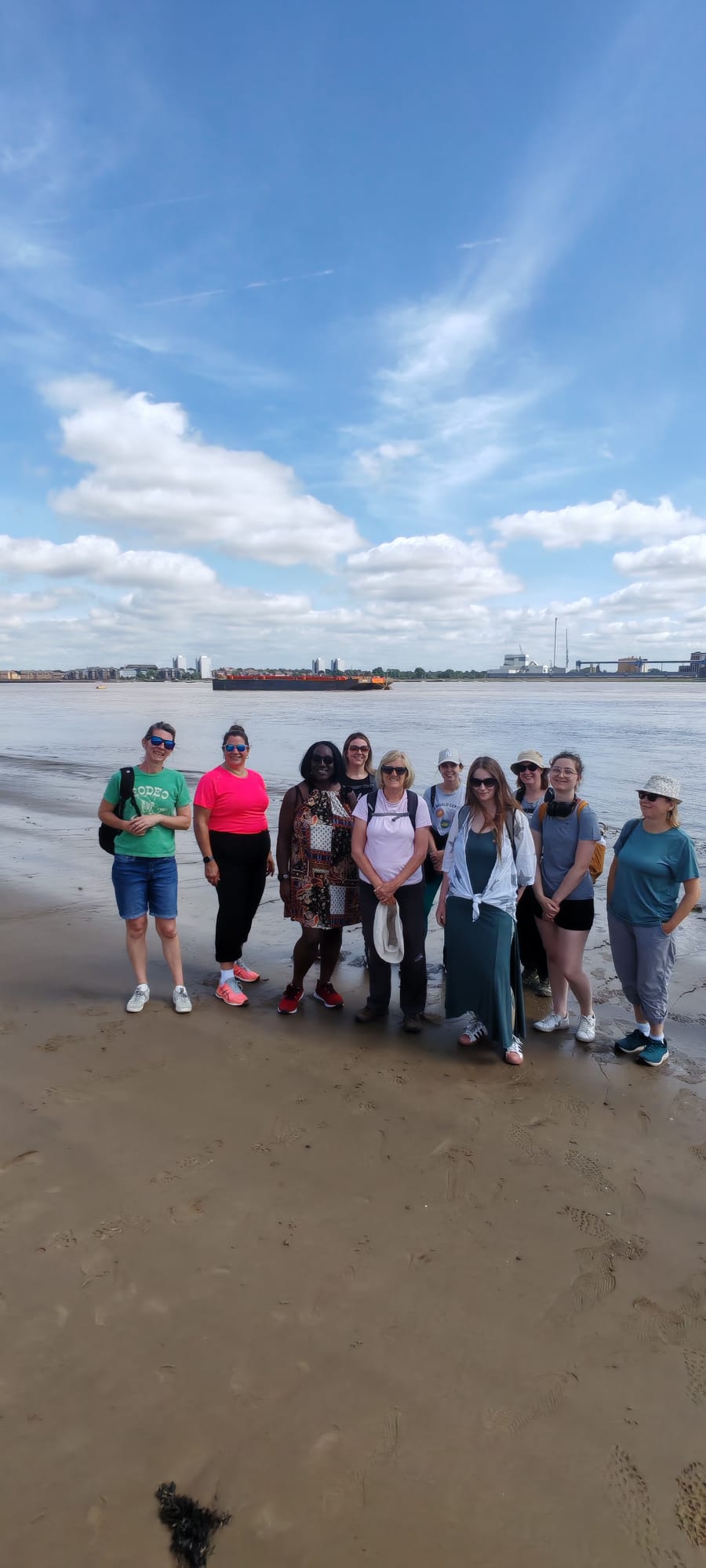 Group of women smiling at the camera standing on Northfleet beach. The River Thames is in the background with a boat in the background.