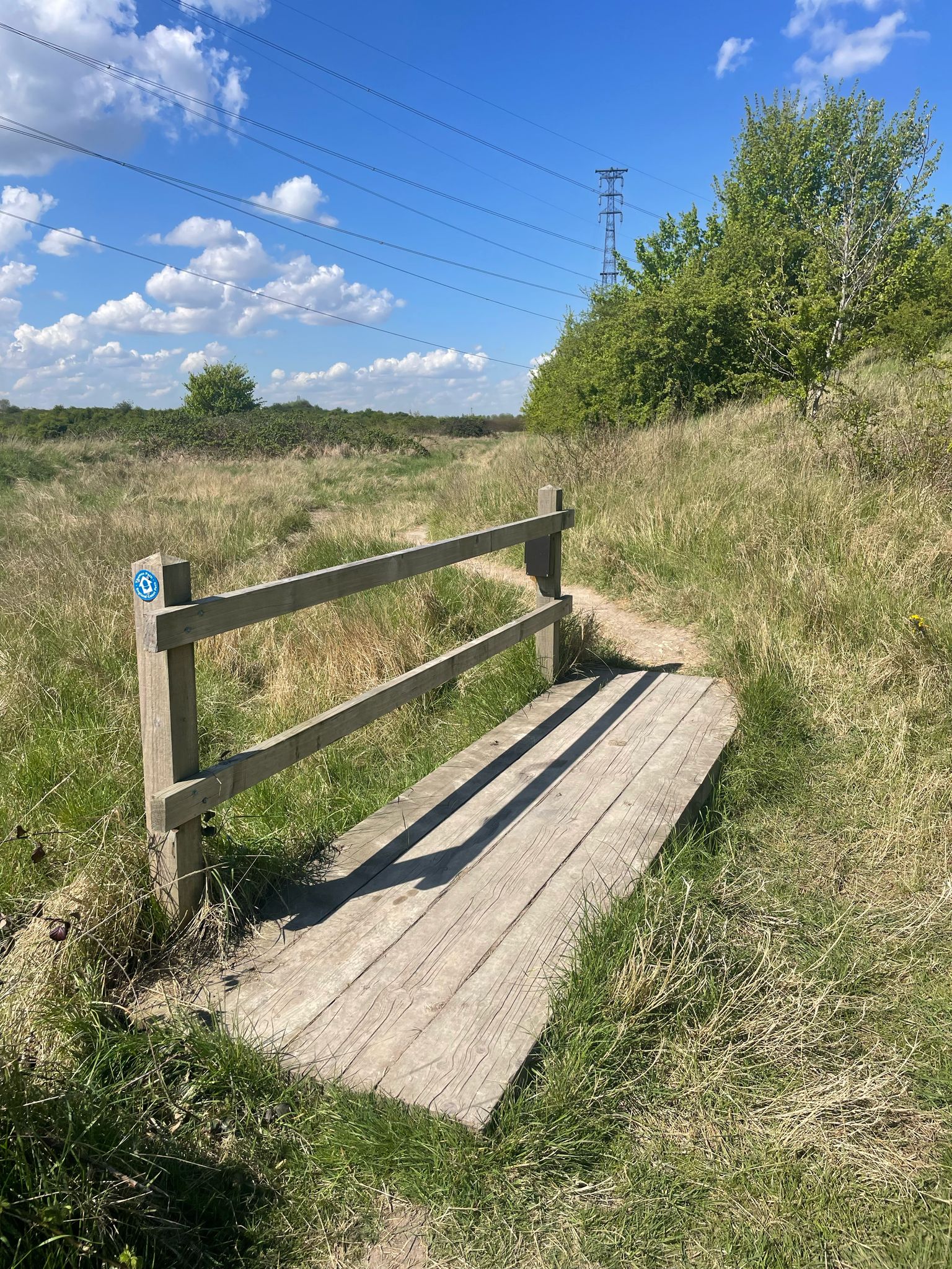 A small wooden footbridge crossing a grassy path in a rural landscape, surrounded by green trees and bushes. The sky is bright blue with scattered clouds, and power lines with an electrical tower are visible in the distance.