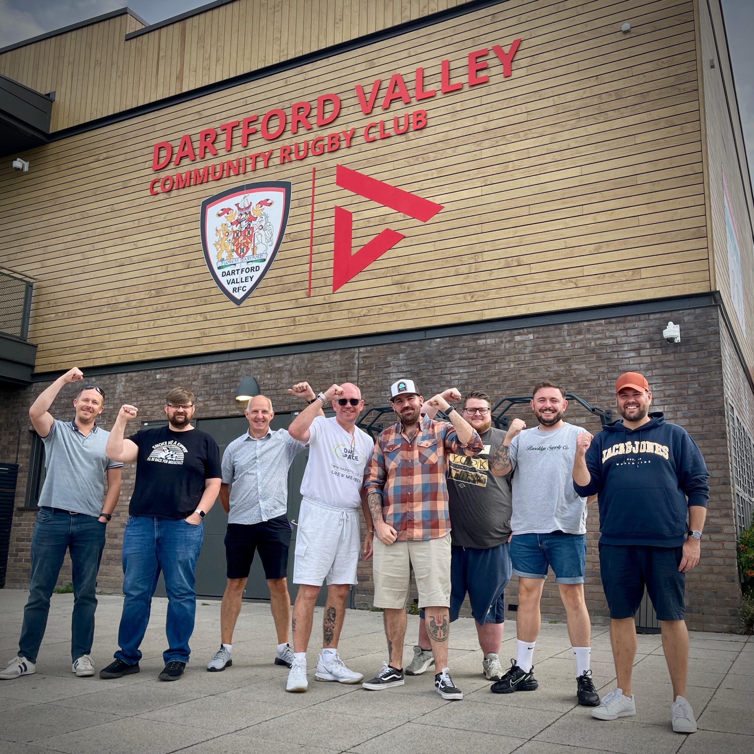 Group of men standing outside Dartford Valley Community Rugby Club. They are smiling at the camera and are raising a fist in the air.
