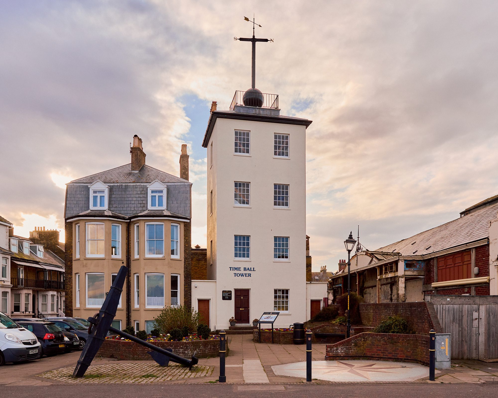Historic Time Ball Tower in a coastal town with a large anchor monument in front, surrounded by garden and adjacent buildings, under soft morning or evening light