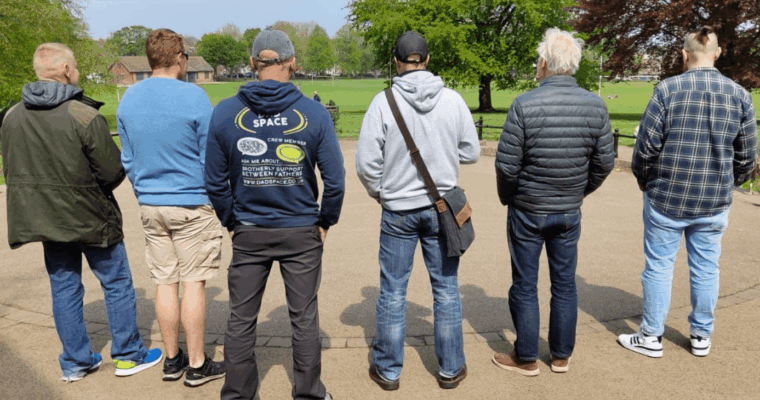 Six men standing in a park with backs turned, one wearing a hoodie promoting fatherhood support