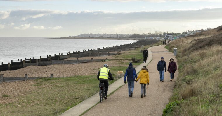 People are walking and cycling along a flat coast path. It is a cold cloudy day and people are wearing coats. The sea is calm and is separated from the path by a shingle beach