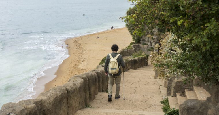 A person with a backpack and walking stick descends stone steps toward a sandy beach with gentle waves and a cloudy sky