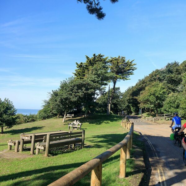 Two people cycling on bikes on a road passing picnic benches with the sea in the background