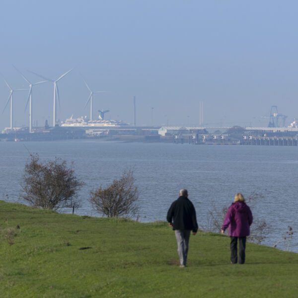 2 people walking along the River Thames with wind turbines and ship in the background