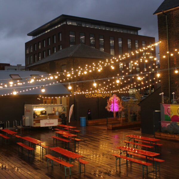 Outdoor seating area in the evening with red picnic benches and strings of lights, Coachworks, Ashford