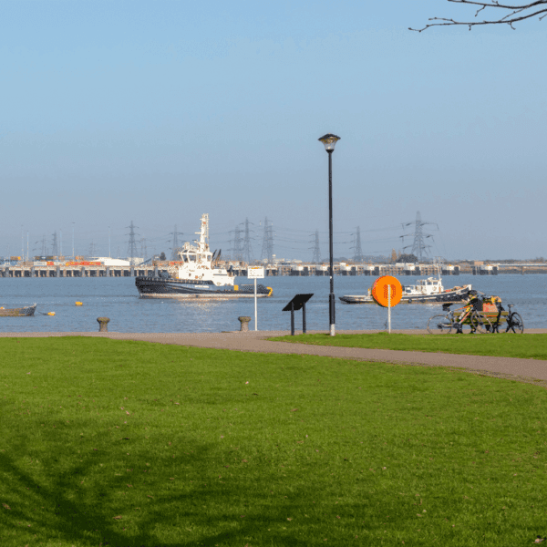 Image shows people walking along Gravesend promenade with grass on one side and the River Thames in the background. There are a boats in the background on the River.