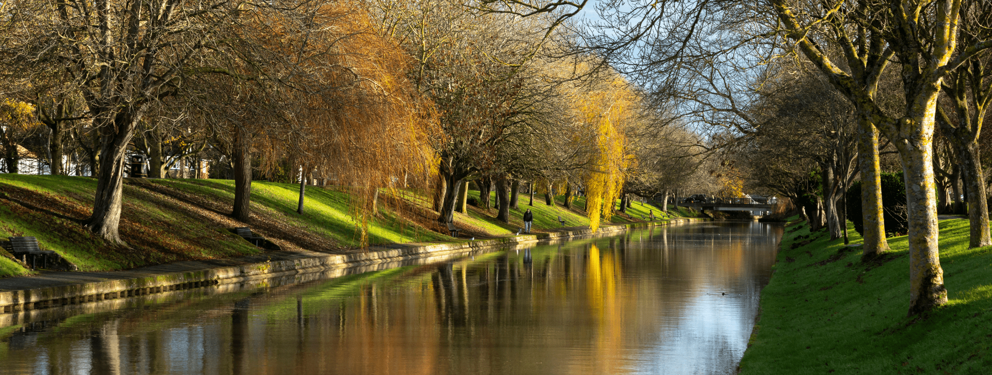 A calm canal lined with bare and golden‑leafed trees reflects the sky and branches in still water. A paved path with benches runs alongside the canal, with a few people walking in the distance and a small bridge visible further ahead.