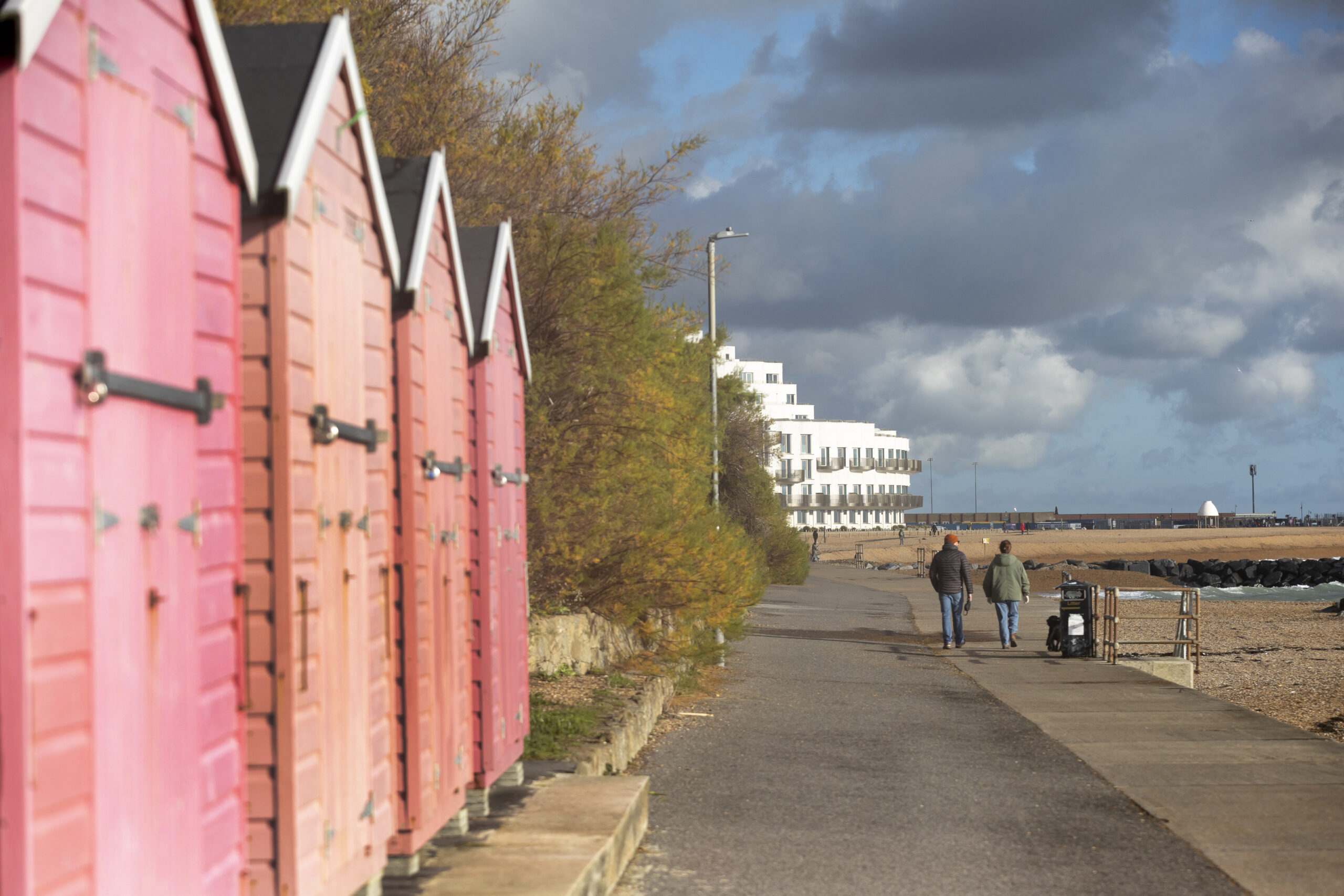 a smooth tarmac path with pink beach huts to the left and people walking in the distance