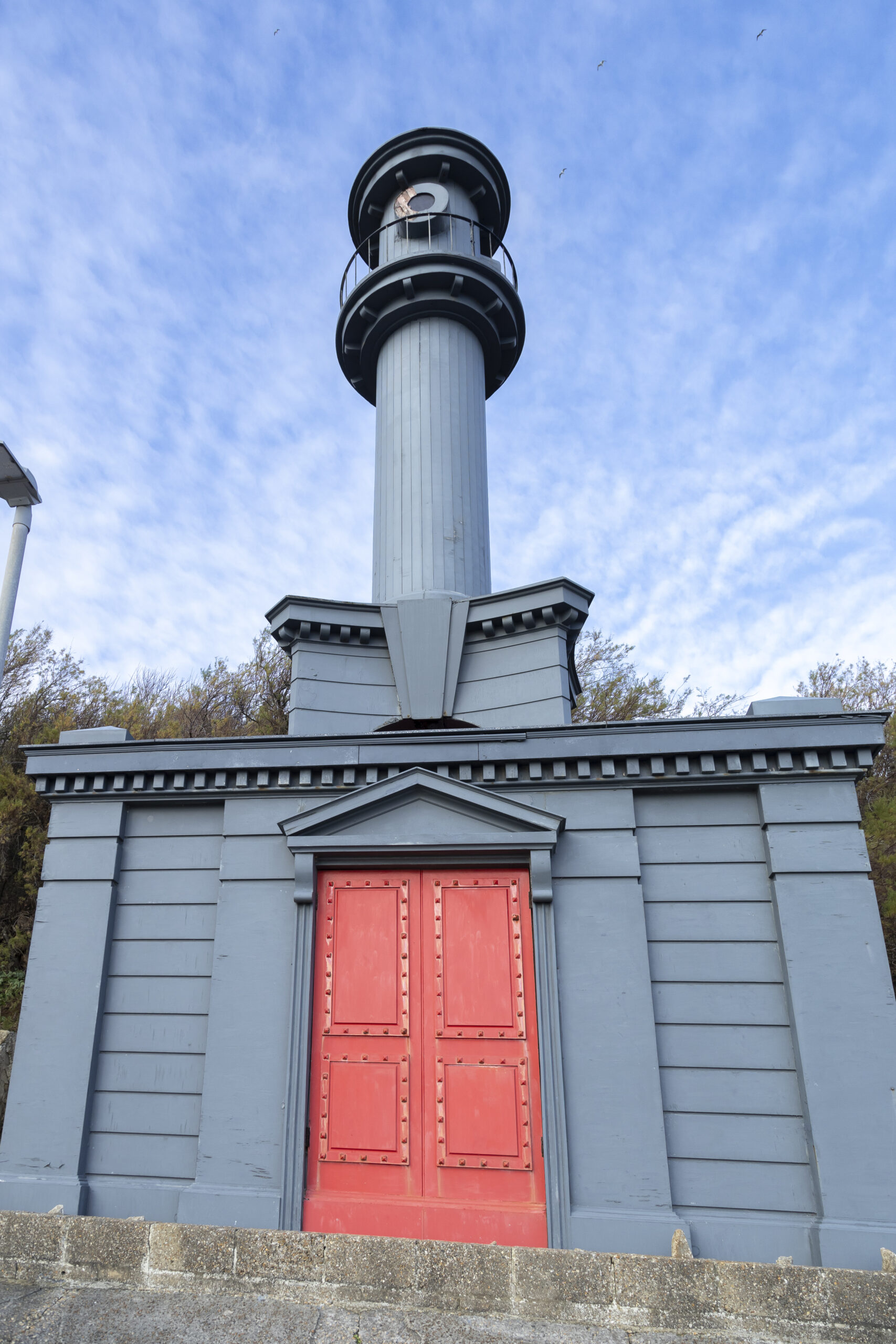 Architectural-style beach hut in Folkestone designed by Pablo Bronstein, resembling a small classical tower with a red door, set among colourful beach huts beside the seafront.