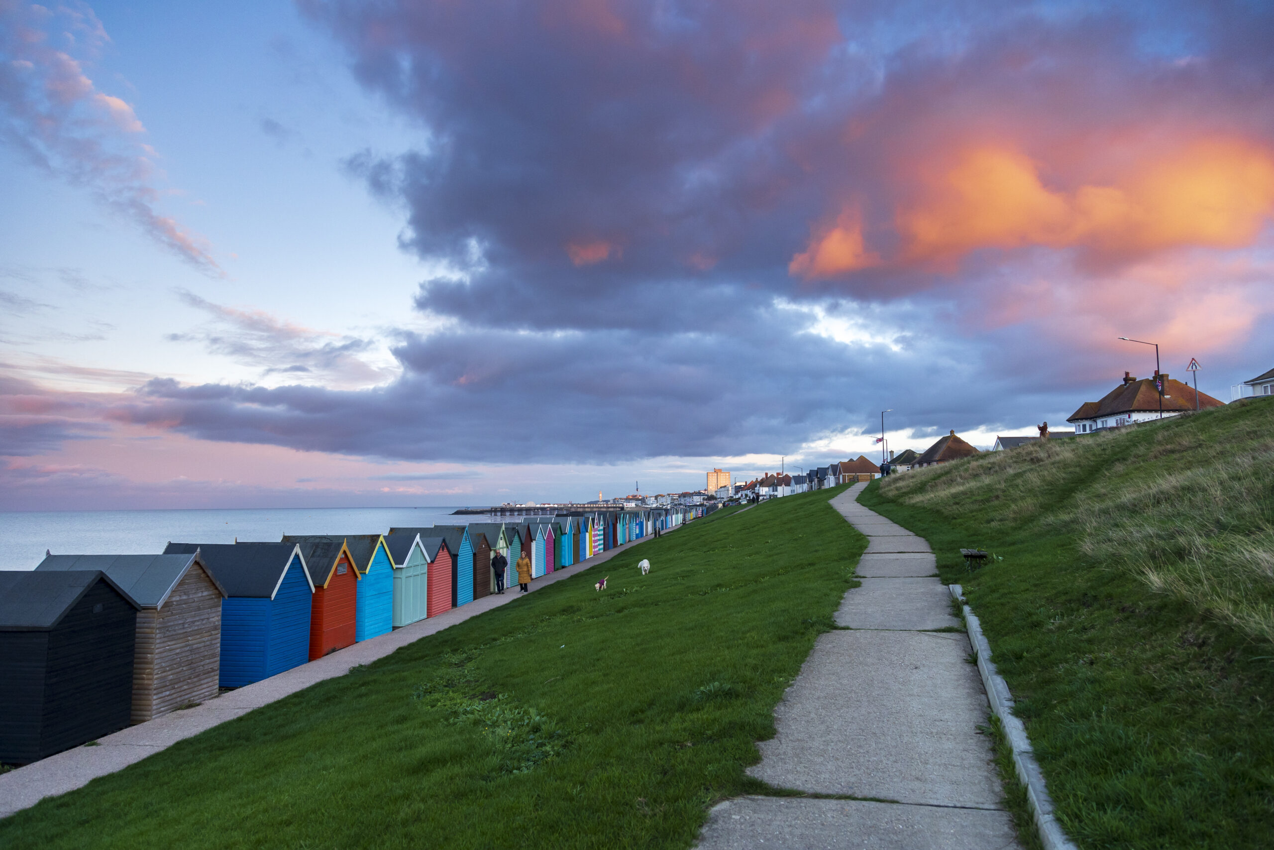 a surfaced path with colourful beach huts to the site. The sky is blue and pink with clouds