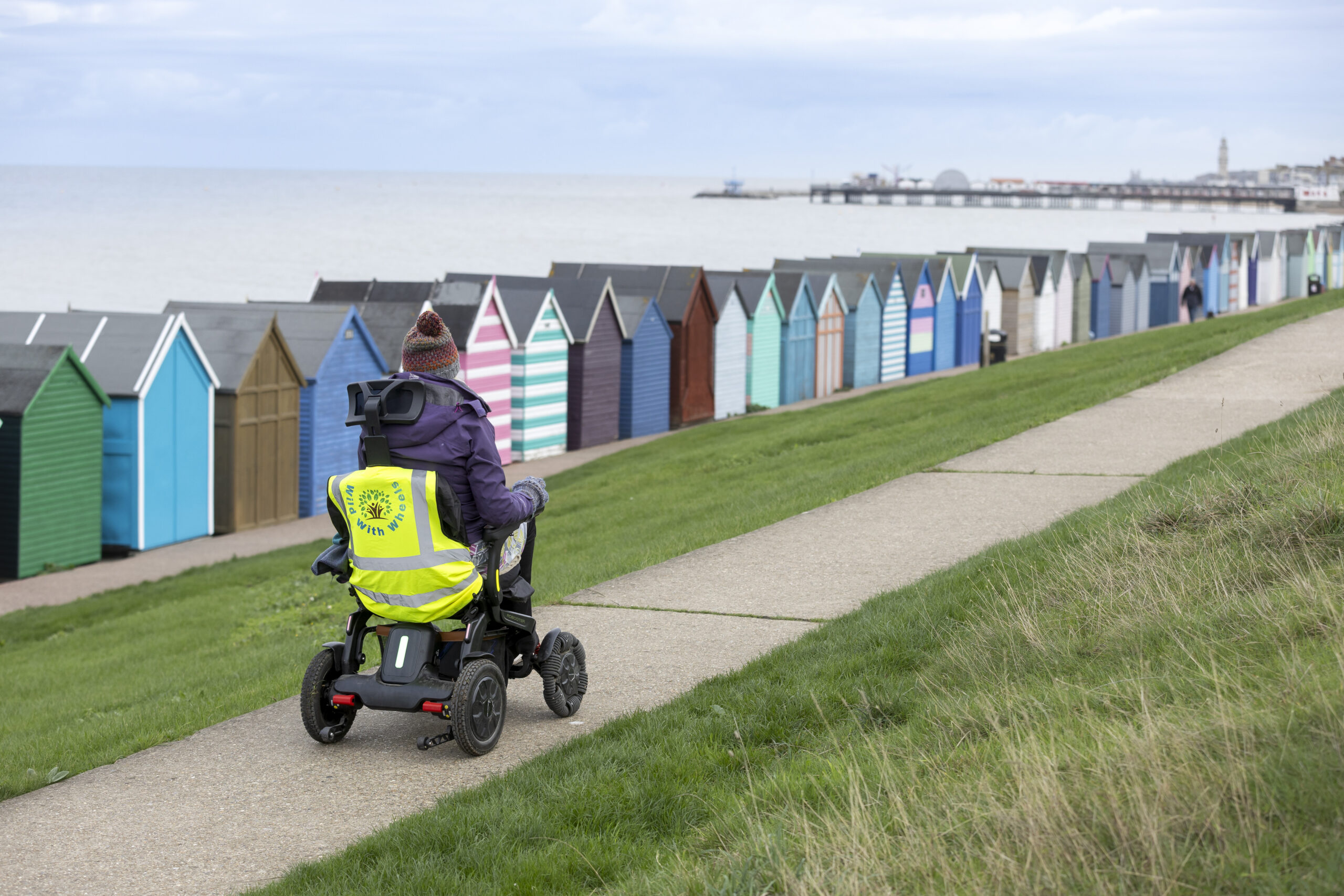 A person wheeling on a motorised wheelchair with colourful beach huts to the side and a view of the sea