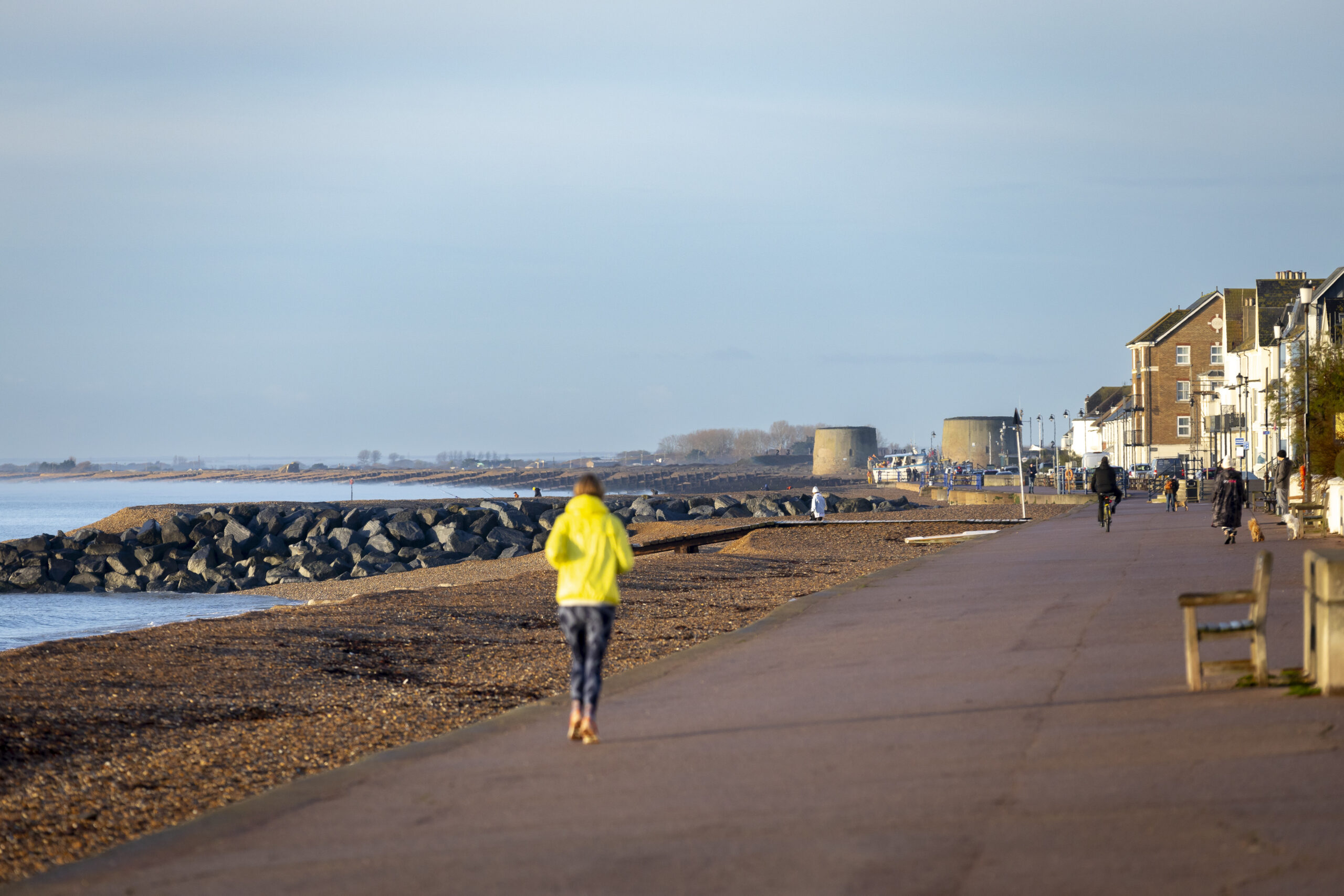 A coastal promenade runs beside a pebble beach, with people walking, cycling and a jogger in a yellow jacket in the foreground. Buildings line the right side of the path, while large rocks form a breakwater on the left. Two round Martello towers stand in the distance under soft daylight.