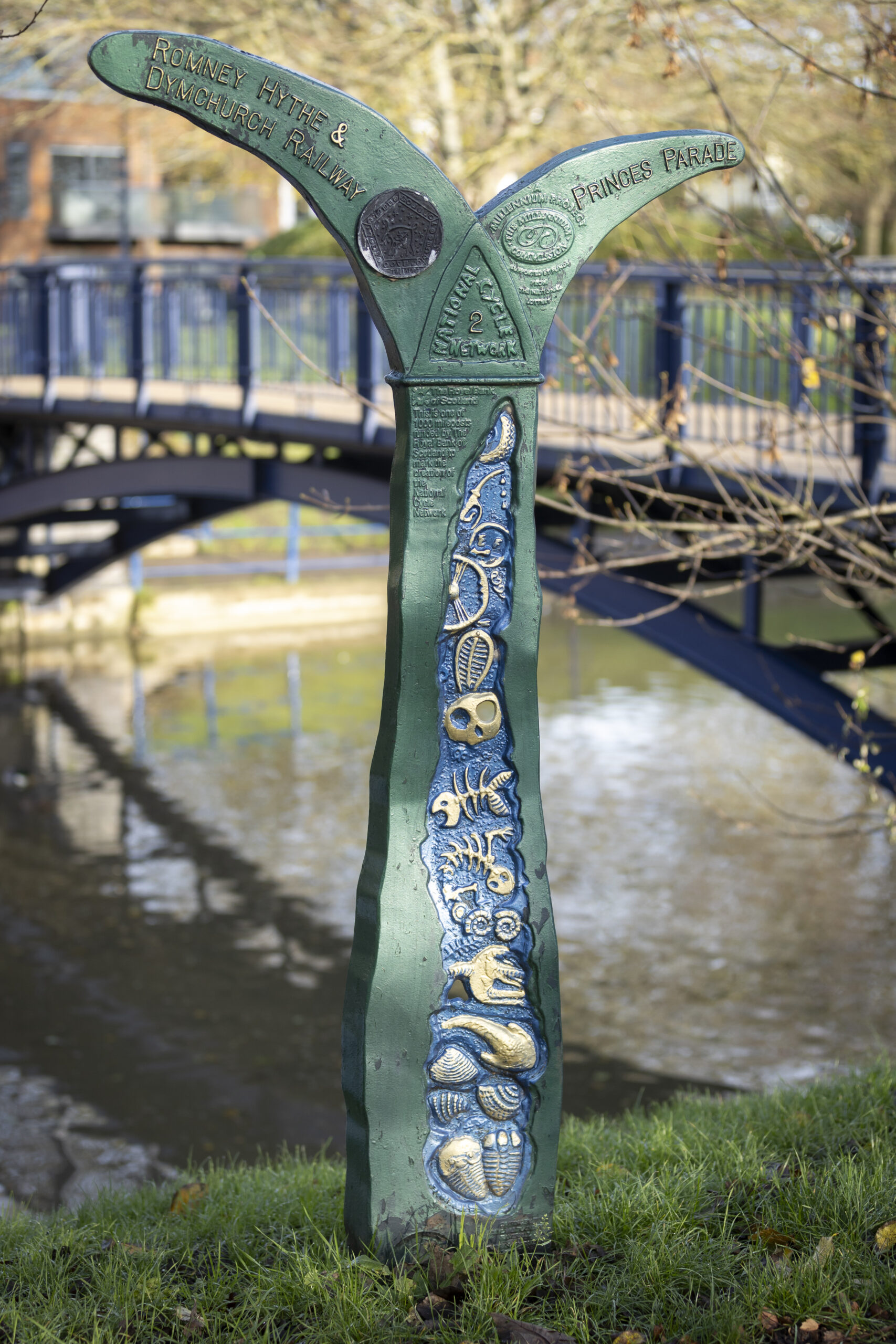 A decorative green metal signpost beside a canal, featuring gold and blue fish and shell motifs. One arm points toward “Romney Hythe & Dymchurch Railway” and the other toward “Princes Parade.” A blue pedestrian bridge crosses the water in the background.