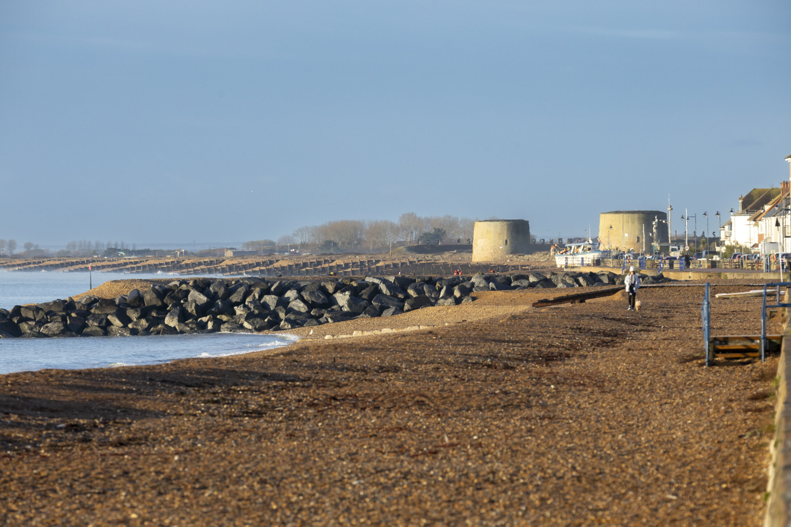 Two large Martello towers stand beside a pebbled beach, with several fishing boats pulled up on the shore. A few people are working near the boats, and trees line the background under warm, low sunlight.