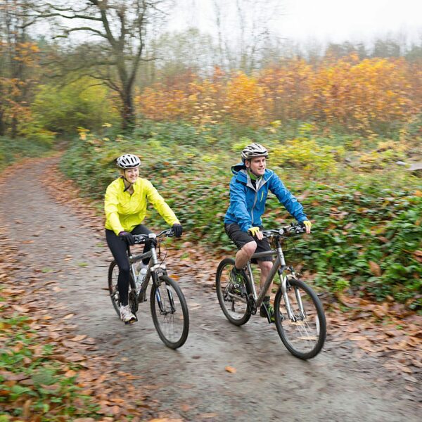 A man wearing a blue coat and a woman wearing a yellow coat cycling on bicycles in Bedgebury.