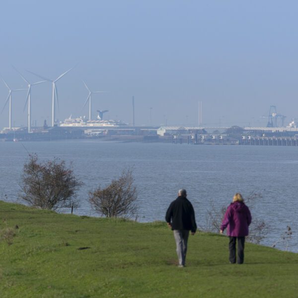 2 people walking along the River Thames with wind turbines and ship in the background