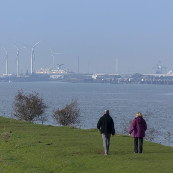 2 people walking along the River Thames with wind turbines and ship in the background