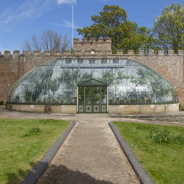 Italianate glasshouse against a brick wall in a garden with a hard surfaced pathway leading to it.