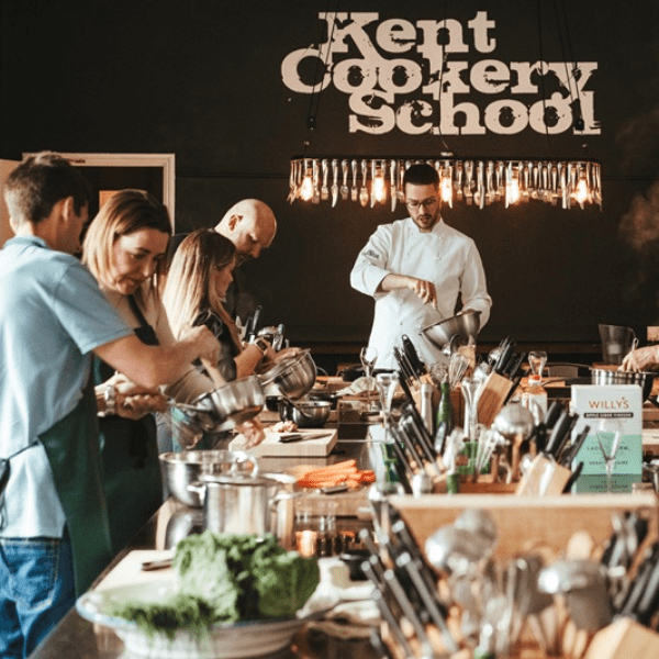 Multiple people cooking at a long metal table being taught by an instructor during a cooking class.