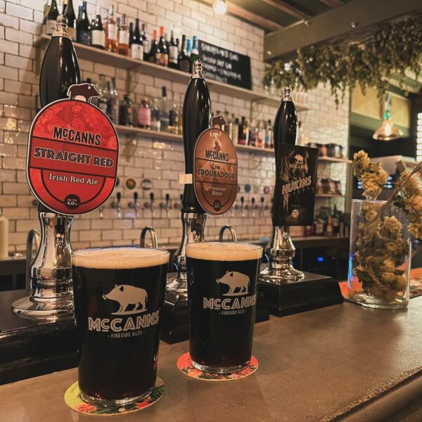 Two pints of beer in McCanns glasses sat on a bar with drinks in the background
