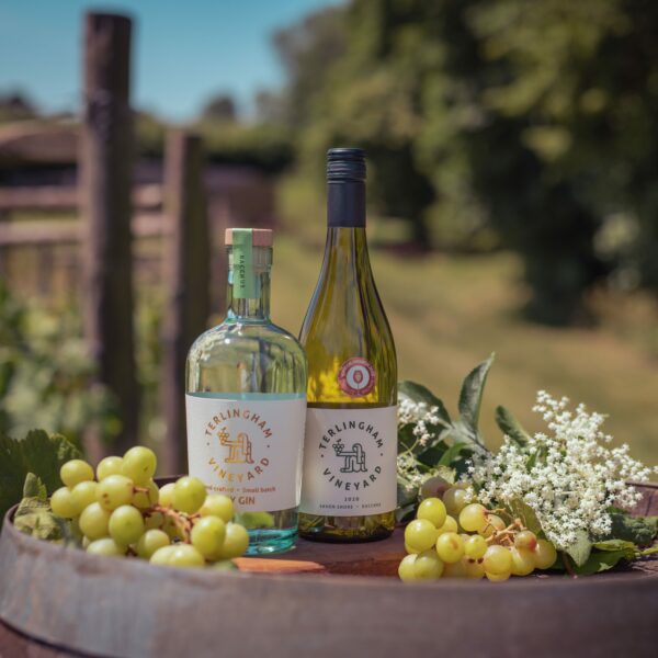 A bottle of Terlingham Vineyard wine next to a bottle of gin on top of a wooden barrel outside with some grapes and white flowers