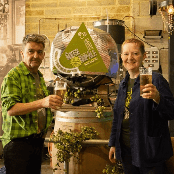 Two people holding pints of ale standing next to a wooden barrel.