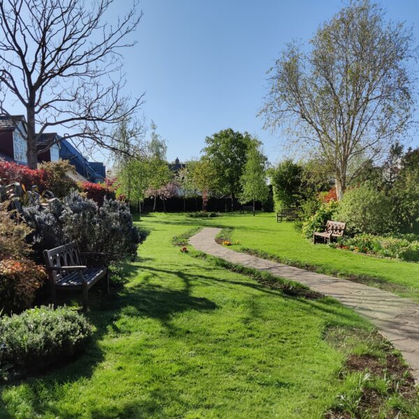 A garden with flower beds, grass lawn, a couple of benches and a winding paved path down the centre.