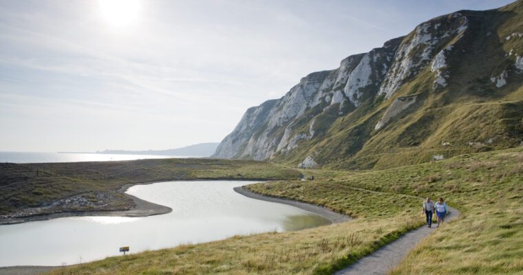 Two people walking along a gravel path between a small lake and white chalk cliffs at Samphire Hoe in Dover, with the sea in the background.