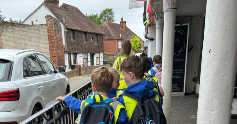 A line of children wearing high-vis jackets and backpacks walking next to a road with a silver car.