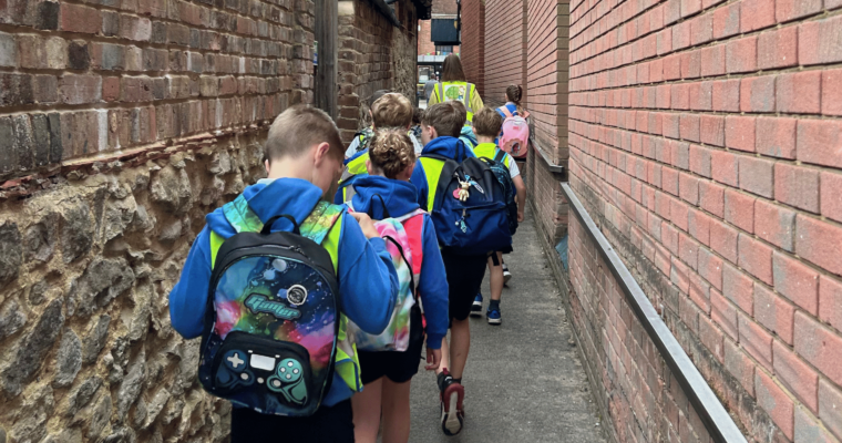 A line of children wearing high-vis jackets and backpacks walking through an alleyway.