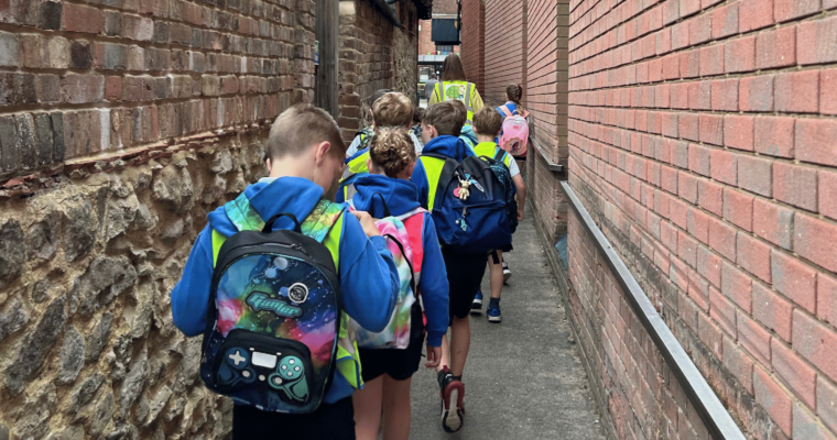 A line of children wearing high-vis jackets and backpacks walking through an alleyway.