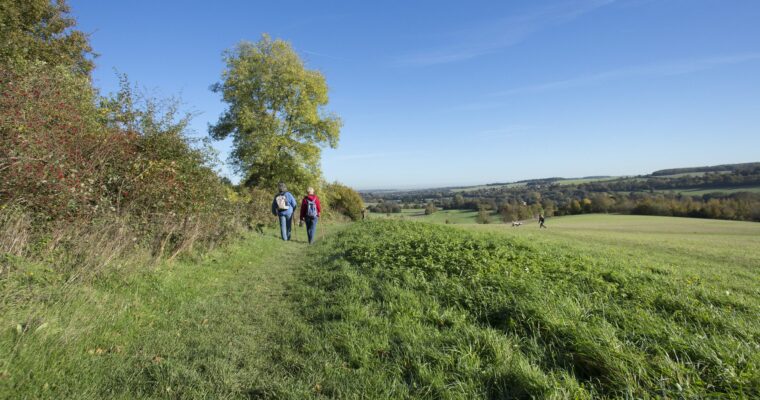 Two people walking on a grassy path with trees to the left and views of Darent Valley to the right, Lullingstone Country Park