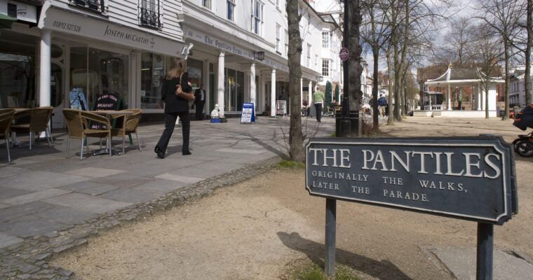A woman walking down a tiled and tree-lined street next to a row of shops. There is a sign for The Pantiles.